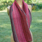 A Striped Cowl with alternating red, pink, purple, and brown vertical stripes is draped over a dress form. The background shows a blurred, grassy outdoor setting with some trees and flowers. This beautiful accessory was crafted using a Yarn & Soul pattern from the Superfine 400 Alpaca Braids.