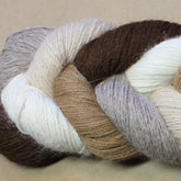 A close-up image of a Yarn & Soul Superfine 400 Alpaca Braid made from multiple skeins of sport weight yarn. The yarn twists together in shades of brown, beige, cream, and gray, creating a visually pleasing gradient of natural colors with excellent stitch definition. The background is a plain, light brown surface.