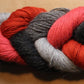 A braided bundle of Yarn & Soul's Superfine 400 Alpaca Braid yarn featuring hues of grey, red, black, and pink. The sport weight strands are tightly interwoven, creating a mixed color pattern against a neutral brown background. This unique blend offers excellent stitch definition for your projects.