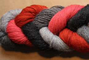 A braided bundle of Yarn & Soul's Superfine 400 Alpaca Braid yarn featuring hues of grey, red, black, and pink. The sport weight strands are tightly interwoven, creating a mixed color pattern against a neutral brown background. This unique blend offers excellent stitch definition for your projects.