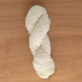 A twisted skein of off-white Stardust Silk Yarn from Henry's Attic is lying on a wooden surface with a natural grain pattern. The yarn appears to be soft yet slightly rough, perfect for creating a gauge swatch that showcases its unique texture and thickness.