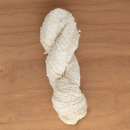 A skein of Stardust Silk Yarn in off-white by Henry's Attic is coiled neatly on a wooden surface, making it an ideal choice for knitters seeking a unique texture. Its slightly curly appearance invites knitting or crocheting and inspires creative weaving projects.