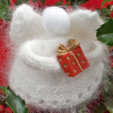 The "Snow Angels" handmade white wool ornament from Halcyon Yarn exudes holiday decor elegance, resembling an angel complete with wings and a fluffy halo, holding a small red gift box adorned with a gold ribbon. The background features green holly leaves and red accents, adding to its festive charm.