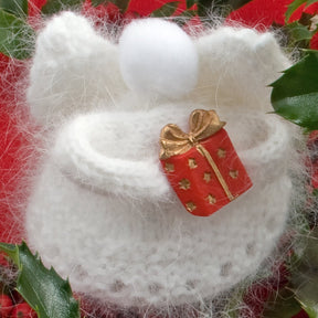 The "Snow Angels" handmade white wool ornament from Halcyon Yarn exudes holiday decor elegance, resembling an angel complete with wings and a fluffy halo, holding a small red gift box adorned with a gold ribbon. The background features green holly leaves and red accents, adding to its festive charm.