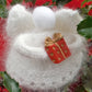 A beautiful ornament from Halcyon Yarn, the "Snow Angels" downloadable pattern features a knitted white angel with fluffy angora wings and a charming pom-pom head, holding a small red gift box adorned with a gold ribbon. This exquisite angel is set against festive green foliage and vibrant red poinsettias, making it perfect for holiday decorating.