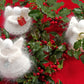 Three "Snow Angels" figurines by Halcyon Yarn, radiating handmade elegance, are arranged amidst lush green holly branches adorned with red berries. The holiday decor features angels holding a snowflake, a gift, and a musical instrument against a backdrop of vibrant red cloth.