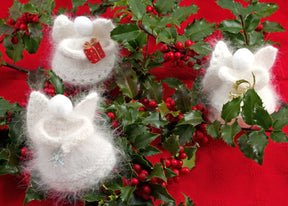 Three "Snow Angels" figurines by Halcyon Yarn, radiating handmade elegance, are arranged amidst lush green holly branches adorned with red berries. The holiday decor features angels holding a snowflake, a gift, and a musical instrument against a backdrop of vibrant red cloth.