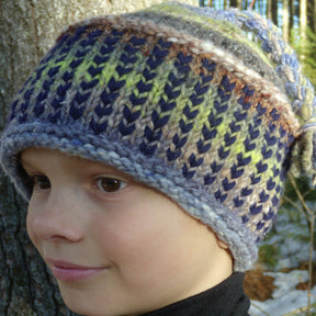 A young person wearing a colorful Alpine Topper Hat from Halcyon Yarn, which features a trek pattern in shades of blue, green, gray, and brown, stands outdoors. The person is smiling slightly and facing left against a backdrop of trees with a hint of snow on the ground.