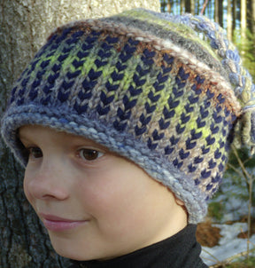 A child wearing a colorful Alpine Topper Hat by Halcyon Yarn stands near a tree trunk. The winter hat, made of merino wool and featuring a zigzag pattern, showcases shades of blue, green, brown, and gray. The background features blurred outdoor scenery with lush green foliage.