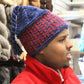 A person with short hair is wearing a Halcyon Yarn Alpine Topper Hat, featuring blue and red colors and a tassel at the top. They are dressed in a red jacket and looking to the right, with shelves of various yarns and knitting supplies in the background.