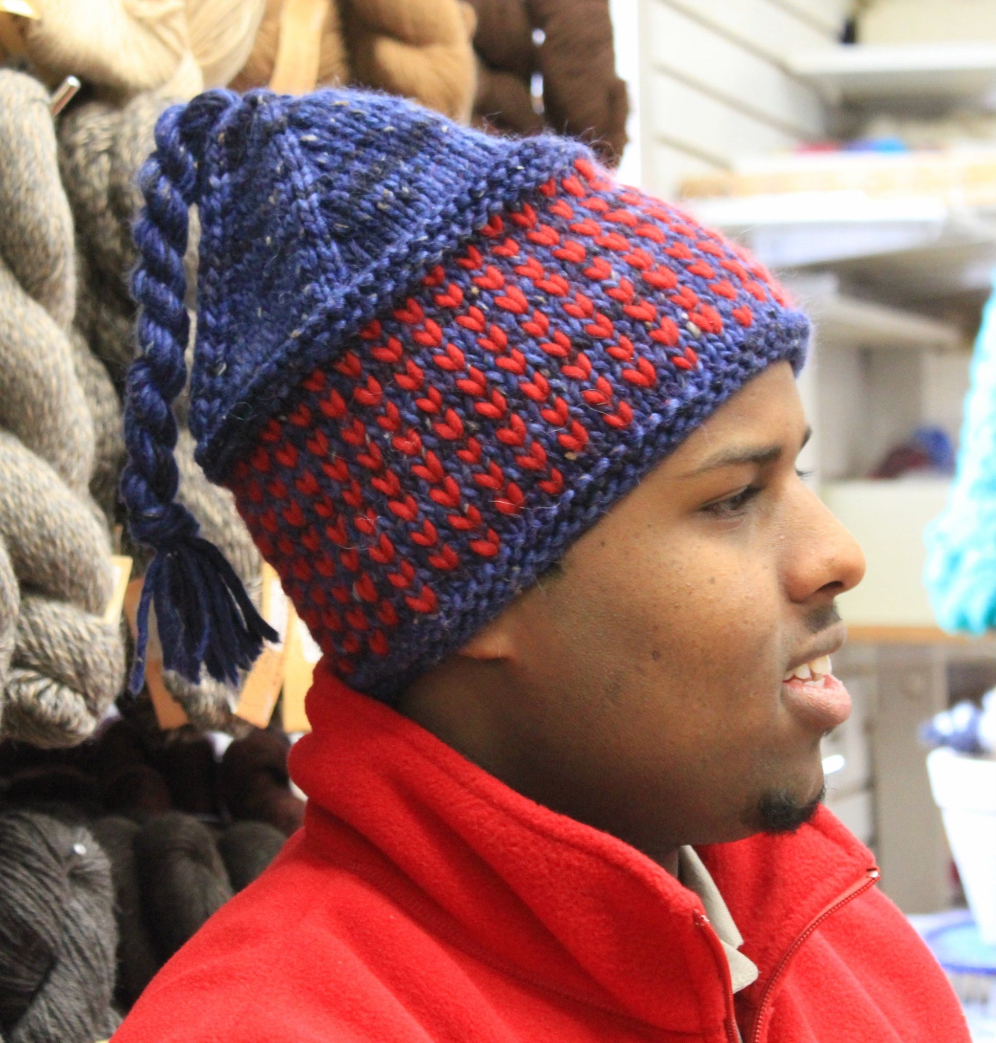 A person with short hair is wearing a Halcyon Yarn Alpine Topper Hat, featuring blue and red colors and a tassel at the top. They are dressed in a red jacket and looking to the right, with shelves of various yarns and knitting supplies in the background.