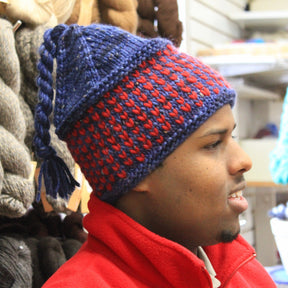 A person wearing the Alpine Topper Hat from Halcyon Yarn in red and blue, with a tassel on top, paired with a red jacket. The individual is in a yarn store, surrounded by various skeins of merino wool in different colors displayed on shelves in the background.