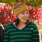 Three women are smiling and wearing colorful striped winter hats, specifically the Alpine Topper Hat by Halcyon Yarn. They are standing outdoors in front of a tree with bright red leaves in the background. The woman on the left is wearing a gray sweater, the middle woman is in a green-striped shirt, and the right woman is dressed in a black shirt.