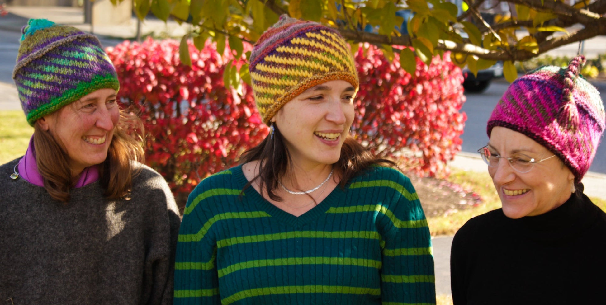 Three women are smiling and wearing colorful striped winter hats, specifically the Alpine Topper Hat by Halcyon Yarn. They are standing outdoors in front of a tree with bright red leaves in the background. The woman on the left is wearing a gray sweater, the middle woman is in a green-striped shirt, and the right woman is dressed in a black shirt.