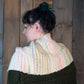 A person with dark hair tied up in a bun is turned to the side, wearing the "Both Sides Now" woven cowl by Halcyon Yarn, which features multicolored knitted patterns in pastel hues and a dark green garment beneath. The background consists of rustic wooden panels, further complementing the fringed cowl that adds an extra touch of elegance.