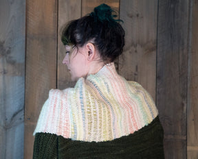 A person with dark hair tied up in a bun is turned to the side, wearing the "Both Sides Now" woven cowl by Halcyon Yarn, which features multicolored knitted patterns in pastel hues and a dark green garment beneath. The background consists of rustic wooden panels, further complementing the fringed cowl that adds an extra touch of elegance.