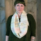 A person with short brown hair and straight bangs is standing against a wooden backdrop, wearing a dark green sweater and the "Both Sides Now, Woven Cowl" by Halcyon Yarn. The individual has visible tattoos on their left forearm and a neutral facial expression.