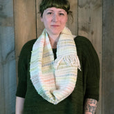 A person with a thoughtful expression stands in front of a wooden wall, wearing a dark green knit sweater and Halcyon Yarn's "Both Sides Now" woven cowl, featuring pastel colors and fringes. They have short, green-tinted hair with blunt bangs and a tattoo visible on their lower right arm.