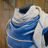 A mannequin showcases the Under the Boardwalk Shawl, available for download from Halcyon Yarn, elegantly draped over its shoulders. The shawl features alternating blue and white stripes in a variety of shades and textures, crafted using garter stitch. Wooden panels in the background add a rustic touch to the image.