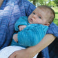 A close-up of a baby being held in an adult's arms outdoors. The baby is wrapped in the colorful and cozy Malabrigo Baby Blanket from Halcyon Yarn and has a serious expression. The person holding the baby is wearing a purple knitted sweater. Green foliage is visible in the background.