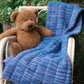 A brown teddy bear is sitting on a white wicker chair with the beautiful Malabrigo Baby Blanket from Halcyon Yarn, knitted in a lovely blue yarn, draped over it. The chair is placed outdoors, surrounded by lush green plants and pink flowers in the background.