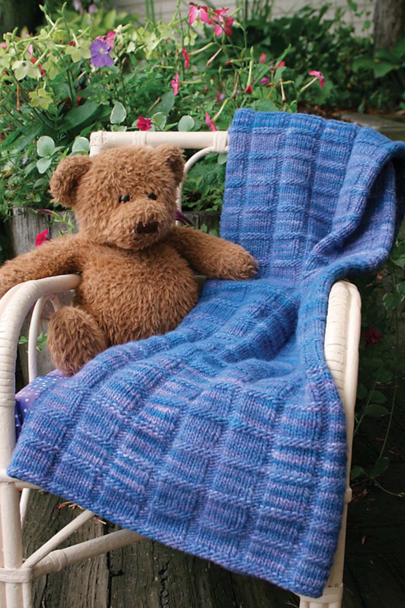 A brown teddy bear is sitting on a white wicker chair with the beautiful Malabrigo Baby Blanket from Halcyon Yarn, knitted in a lovely blue yarn, draped over it. The chair is placed outdoors, surrounded by lush green plants and pink flowers in the background.