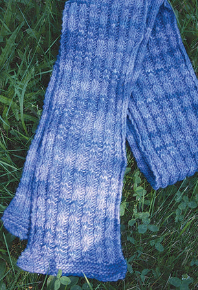 A thick, hand-knitted Malabrigo Mock Cable Scarf from Halcyon Yarn rests on lush green grass. Crafted with Malabrigo worsted weight yarn, it features a textured pattern with alternating shades of blue, providing a cozy and warm appearance.