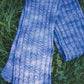 On a bed of grass, the Malabrigo Mock Cable Scarf from Halcyon Yarn is draped elegantly. This reversible accessory features vertical patterns in a knitted, intricate design crafted from hand-dyed yarn. The shades of blue create a stunning gradient effect that contrasts beautifully with the lush green background.