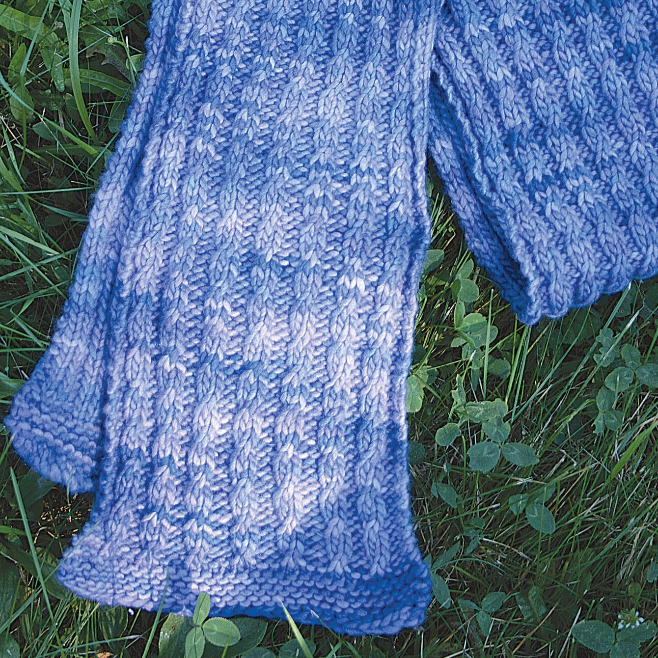 The Malabrigo Mock Cable Scarf by Halcyon Yarn, a cozy and hand-knitted piece in a stunning shade of blue, is made from luxurious worsted weight yarn. It is beautifully laid on green grass adorned with clover leaves, highlighting the scarf's rich texture and exquisite craftsmanship against the natural outdoor setting.