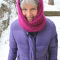 A person with short gray hair stands outdoors in a snowy setting, smiling at the camera. They are wearing a bright pink Halcyon Yarn Rippling Ringlet Infinity Cowl wrapped around their neck and head, a purple winter jacket, and black gloves.