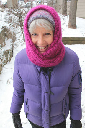 A person with short gray hair stands outdoors in a snowy setting, smiling at the camera. They are wearing a bright pink Halcyon Yarn Rippling Ringlet Infinity Cowl wrapped around their neck and head, a purple winter jacket, and black gloves.