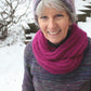 An older woman with gray hair smiles while standing outside in the snow. She is wearing a purple and pink knit beanie, a bright pink Halcyon Yarn Rippling Ringlet Infinity Cowl around her neck, and a multicolored sweater. Leafless trees and snow-covered ground are visible in the background, capturing a serene moment for knitters to appreciate.