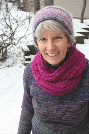 An older woman with gray hair smiles while standing outside in the snow. She is wearing a purple and pink knit beanie, a bright pink Halcyon Yarn Rippling Ringlet Infinity Cowl around her neck, and a multicolored sweater. Leafless trees and snow-covered ground are visible in the background, capturing a serene moment for knitters to appreciate.