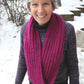 A smiling individual stands outside in a snowy landscape, proudly displaying their Rippling Ringlet Infinity Cowl by Halcyon Yarn—a piece that both novice and seasoned knitters would appreciate. They sport a striped multicolored knit hat, a dark sweater, and this thick, magenta knitted scarf. In the background, small trees and a building can be seen.