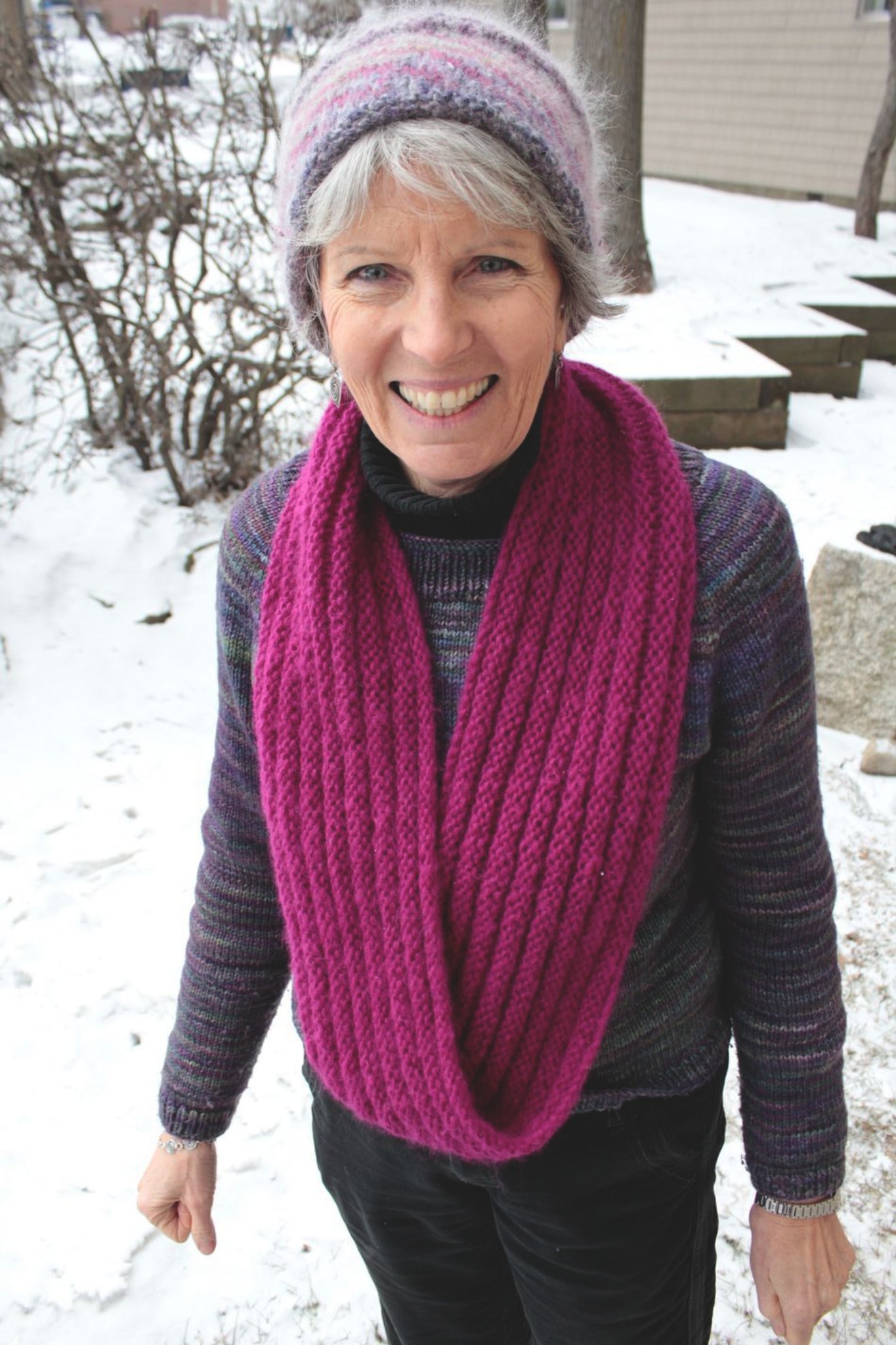 A smiling individual stands outside in a snowy landscape, proudly displaying their Rippling Ringlet Infinity Cowl by Halcyon Yarn—a piece that both novice and seasoned knitters would appreciate. They sport a striped multicolored knit hat, a dark sweater, and this thick, magenta knitted scarf. In the background, small trees and a building can be seen.