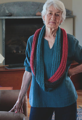 An elderly woman with white hair stands confidently in a living room, wearing a blue blouse and a vibrant Halcyon Yarn Rippling Ringlet Infinity Cowl made by local knitters. Her hand rests on the back of a chair, with the cozy atmosphere enhanced by the warm glow of the fireplace in the background.