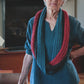 An elderly woman with short white hair is standing in a room, wearing a blue blouse and a Halcyon Yarn Rippling Ringlet Infinity Cowl, featuring red and blue sections. She has one hand on a chair and the other on her hip, smiling slightly. The cozy fireplace in the background underscores her knitting expertise with this downloadable pattern that is perfect for both beginners and experienced knitters.