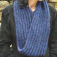 A person is wearing a dark, ribbed coat and the Halcyon Yarn Rippling Ringlet Infinity Cowl knitted with a blend of blue and purple yarn. The cowl is draped loosely around their neck, set against a stone wall background.