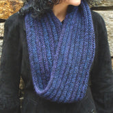 A person with dark curly hair wears a large, chunky knit Halcyon Yarn Rippling Ringlet Infinity Cowl, download, in shades of blue and purple. The scarf is wrapped loosely around their neck. They are dressed in a dark coat, and the background features a stone wall. It's an ideal project for experienced knitters.