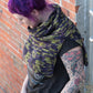 A person with vibrant purple hair stands against a brick wall, wearing the "50,000 Spaceships, Crocheted Ponchette" from Halcyon Yarn. The ponchette features dark purple and green patterns made from Malabrigo Merino Worsted yarn. The individual has several colorful tattoos on their arm and is dressed in a dark T-shirt. They look down with a hint of a smile visible on their face.