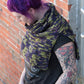 A person with vibrant purple hair stands while looking down. They are wearing a black graphic t-shirt and a green and purple crocheted layering piece, specifically the "50,000 Spaceships" Crocheted Ponchette from Halcyon Yarn, made from Malabrigo Merino Worsted. Their arm is adorned with various colorful tattoos, and they are positioned against a brick wall background.