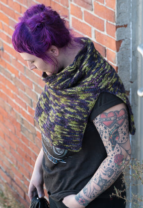 A person with vibrant purple hair stands while looking down. They are wearing a black graphic t-shirt and a green and purple crocheted layering piece, specifically the "50,000 Spaceships" Crocheted Ponchette from Halcyon Yarn, made from Malabrigo Merino Worsted. Their arm is adorned with various colorful tattoos, and they are positioned against a brick wall background.