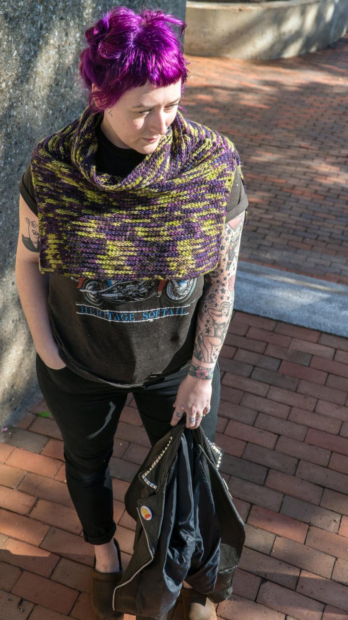 A person with short, bright purple hair stands outdoors on a brick pathway. They are wearing a black T-shirt, black pants, and brown shoes along with the 50,000 Spaceships Crocheted Ponchette by Halcyon Yarn. One hand is in their pocket, and the other holds a black jacket. Tattoo sleeves are visible on both arms.