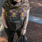 A person with bright purple hair stands on a brick pathway, wearing a dark T-shirt with a graphic design, black pants, and the "50,000 Spaceships" Crocheted Ponchette from Halcyon Yarn. They are holding a black jacket in one hand and have several tattoos on both arms.