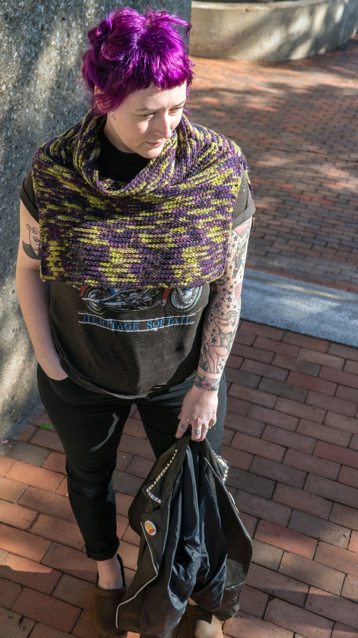 A person with bright purple hair stands on a brick pathway, wearing a dark T-shirt with a graphic design, black pants, and the "50,000 Spaceships" Crocheted Ponchette from Halcyon Yarn. They are holding a black jacket in one hand and have several tattoos on both arms.