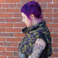 A person with purple hair and visible tattoos stands against a brick wall, looking down to the left. They are wearing a vibrantly colored ponchette called "50,000 Spaceships," crocheted from Malabrigo Merino Worsted yarn in shades of green, yellow, and purple by Halcyon Yarn, paired with a black T-shirt. The image captures a side profile of their upper body.