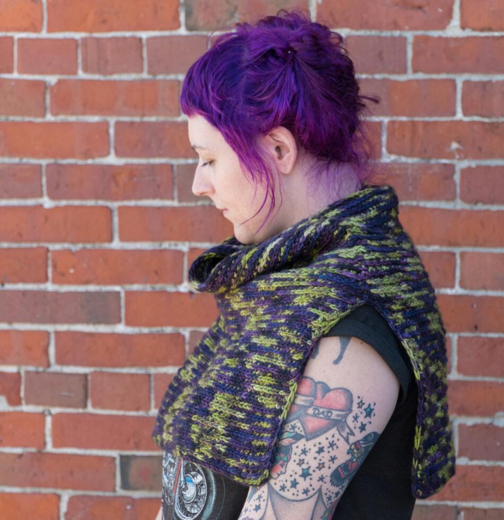 A person with purple hair and visible tattoos stands against a brick wall, looking down to the left. They are wearing a vibrantly colored ponchette called "50,000 Spaceships," crocheted from Malabrigo Merino Worsted yarn in shades of green, yellow, and purple by Halcyon Yarn, paired with a black T-shirt. The image captures a side profile of their upper body.