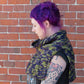 A person with vibrant purple hair stands in profile against a brick wall, wearing a sleeveless black top that showcases several colorful tattoos on their arm. They also sport a versatile accessory—a multicolored ponchette made from the "50,000 Spaceships" pattern by Halcyon Yarn, featuring shades of green, purple, and black.