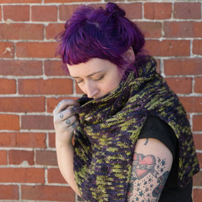 A person with vibrant purple hair is standing against a brick wall. They are wearing a black top and the "50,000 Spaceships, Crocheted Ponchette" from Halcyon Yarn, crafted from shades of green and purple worsted weight yarn. With tattoos on their arms, they gently hold the ponchette close to their face, looking down.