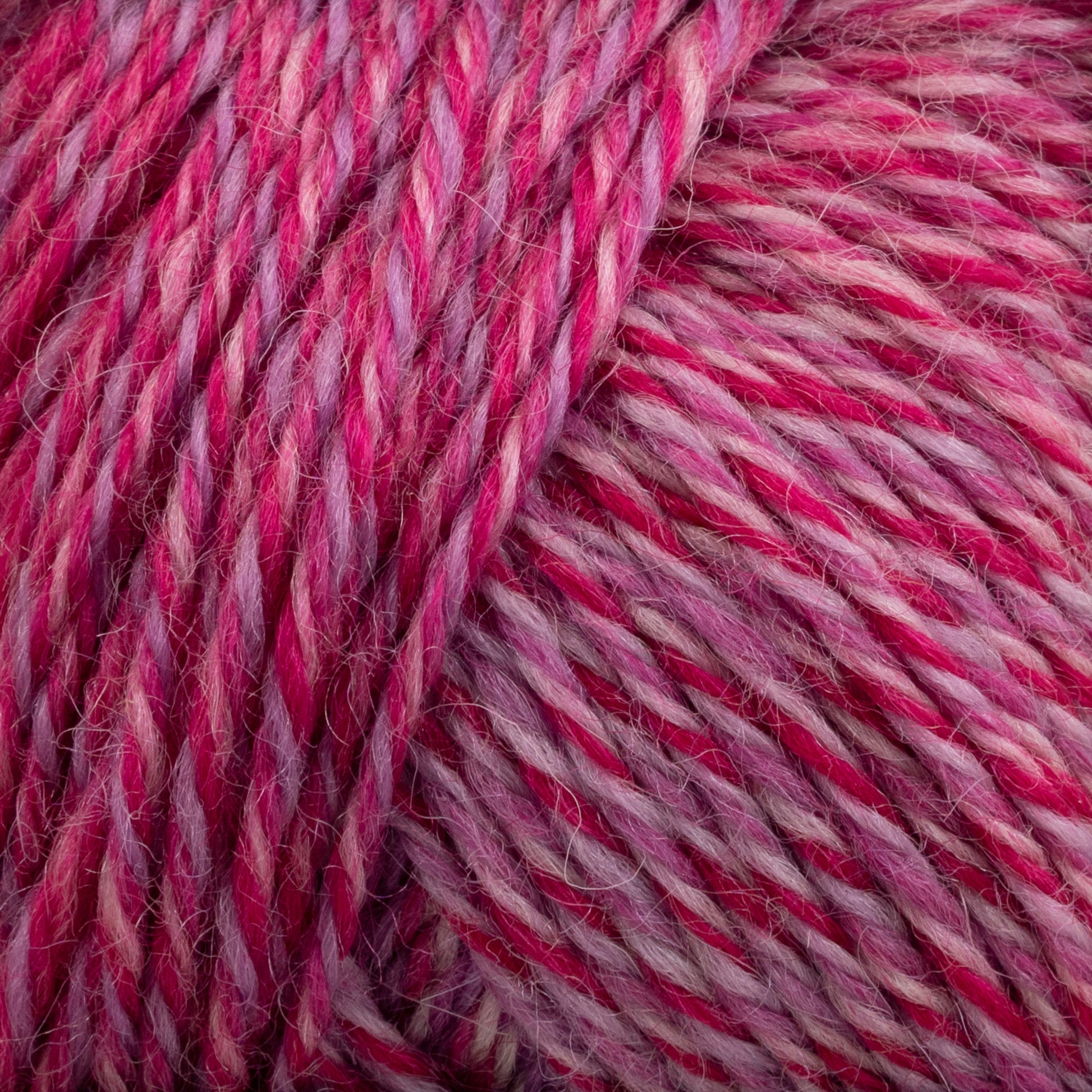 Close-up image of Painted Sky Wool by Knitting Fever / Euro, featuring a pink and purple variegated yarn made from Australian wool. The fibers are tightly twisted together, showcasing a mix of vibrant and subdued hues with long gradient color changes, creating a textured and colorful appearance. This versatile yarn appears soft and suitable for knitting or crocheting projects.